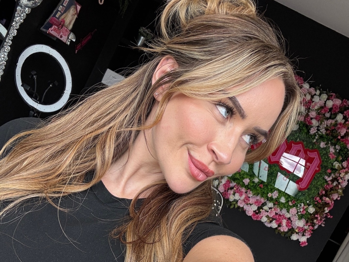 Alice in her clinic, with the ring light and the neon sign on the flower wall behind her
